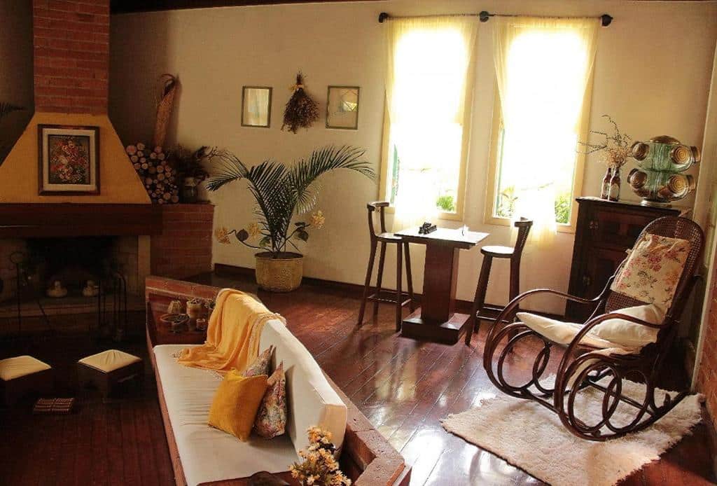 sala rústica com sofá, cadeira de balanço, janela com cortina e lareira tudo em detalhes amarelos no Refúgio Pasárgada Guest House, uma das pousadas em Mairiporã