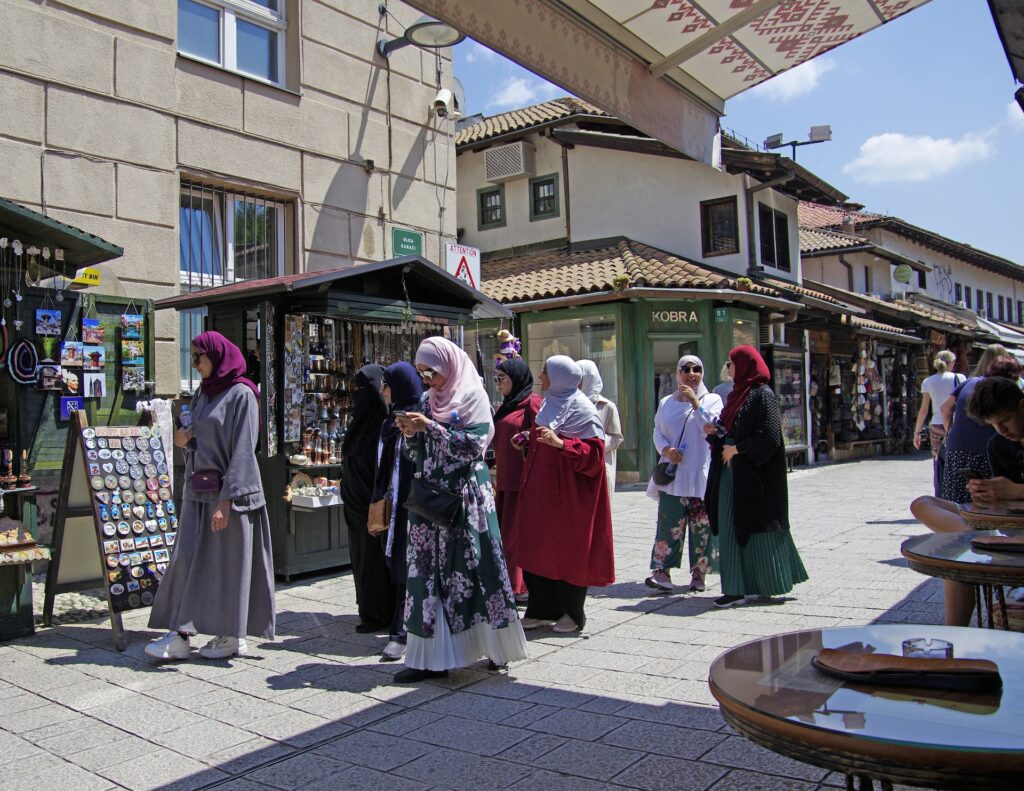 Mulheres andando com turbantes na cabeça, mexendo no celular e conversando, num calçadão de Sarajevo, capital do país, com alguns pontos de comércios e mesas redondas de um restaurante em volta