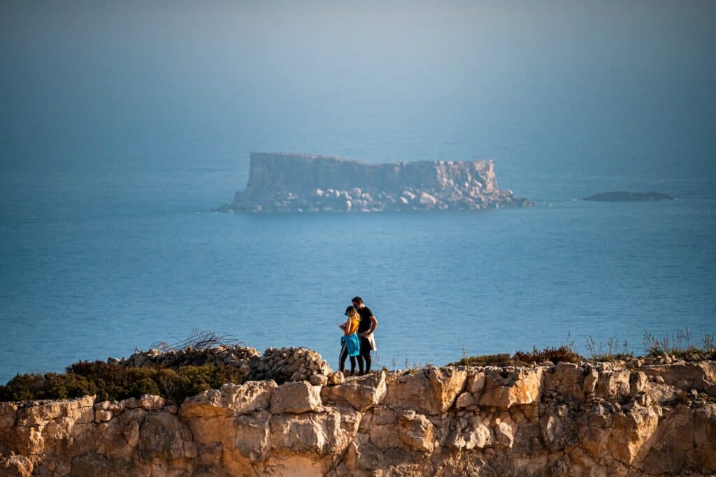 Homem e mulher em um Mirante de pedras, olhando algo que está nas mãos da menina, com vista do mar e de uma pequena ilha no meio, num dia ensolarado