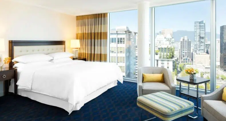 Quarto no Sheraton Vancouver Wall Centre com uma cama grande, poltrona, pufe e janelas do chão ao teto com vista para a paisagem urbana, para representar hotéis em Vancouver.