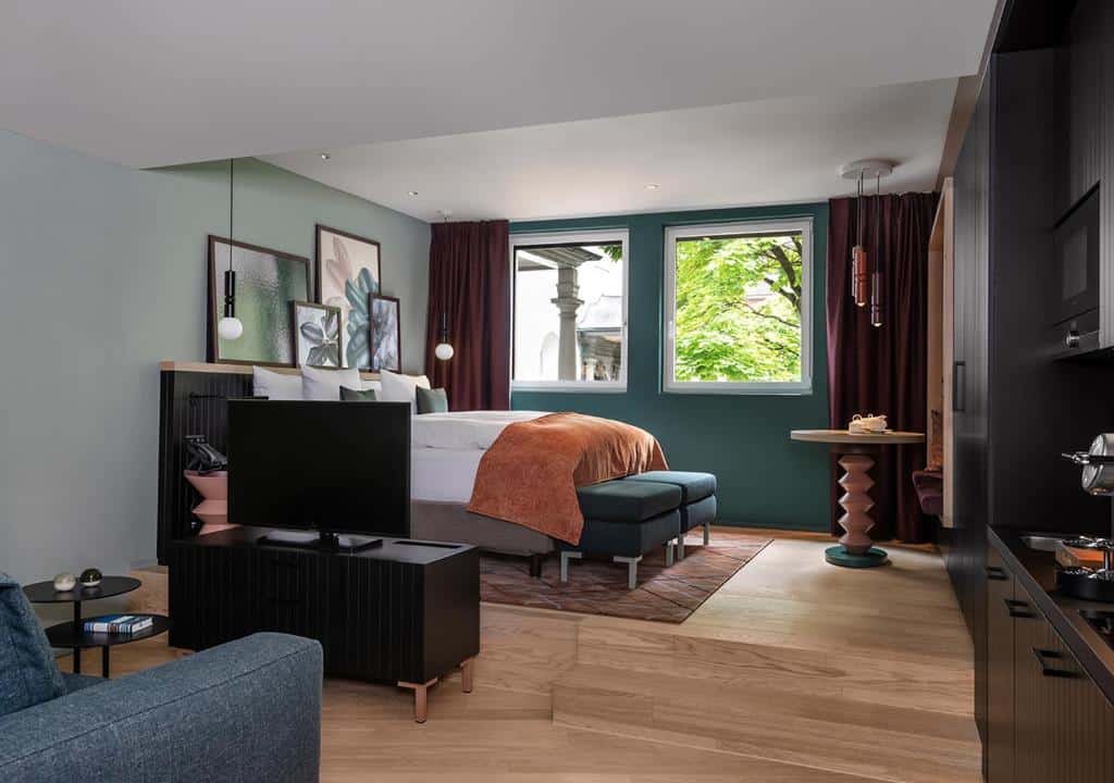 Quarto Grand deluxe do Sorell Hotel St. Peter, de 38 m², com cama de casal macia, quadros pendurados, TV, sofá, mini cozinha com móveis pretos e janela mostrando uma árvore verde