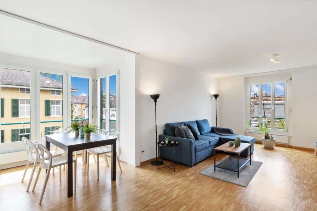 Sala de estar com pequena copa integrada do Spacious Central Apartments, com janelas de vidro mostrando prédios das redondezas e céu azul, além de um sofá azul turquesa, pequena mesa de centro e mesa com quatro cadeiras