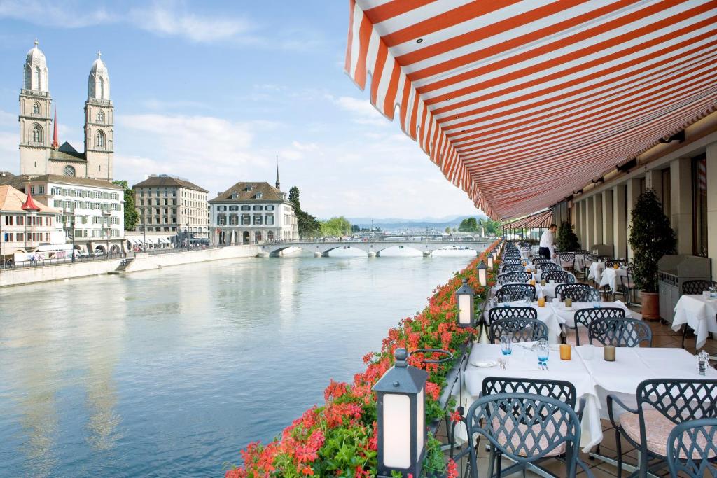 Varanda e restaurante do Storchen Zürich Boutique Hotel, em Zurique, com mesas enfileiradas e um garçom no meio delas, usando calça preta e blusa branca, tendo um rio cristalino ao lado, e uma ponte dando acesso a uma região com construções tradicionais da cidade