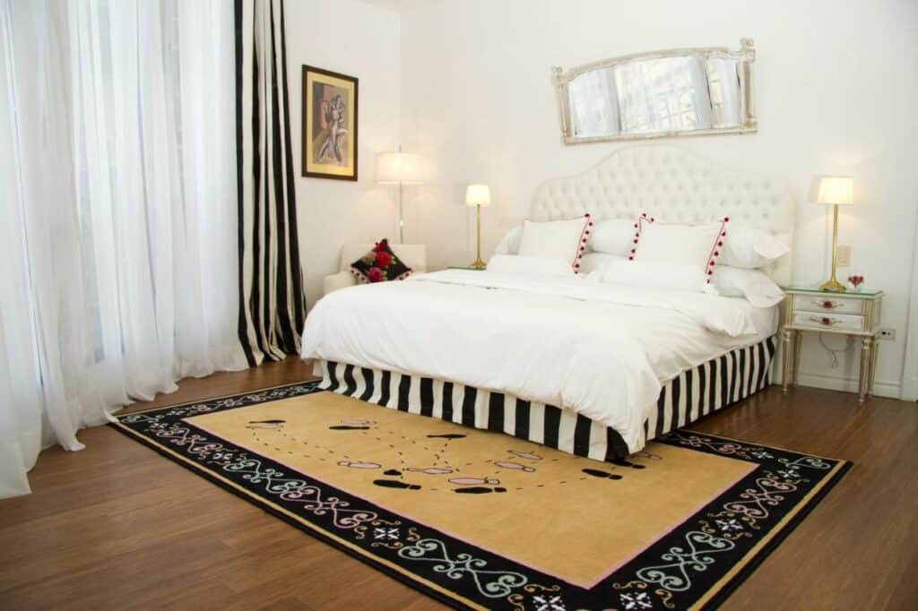 Quarto grande do Tango de Mayo Hotel com uma cama de casal, duas mesinhas de cabeceira com abajures, uma poltrona, uma janela com cortinas, chão de madeira e um tapete