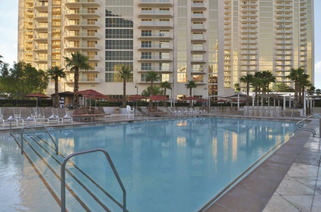 Piscina ampla do The Signature at MGM Grand - All Suites de frente para as duas torres do hotel e, ao redor da piscina, há espreguiçadeiras beges. Há barras de apoio e escadas para entrar na piscina.