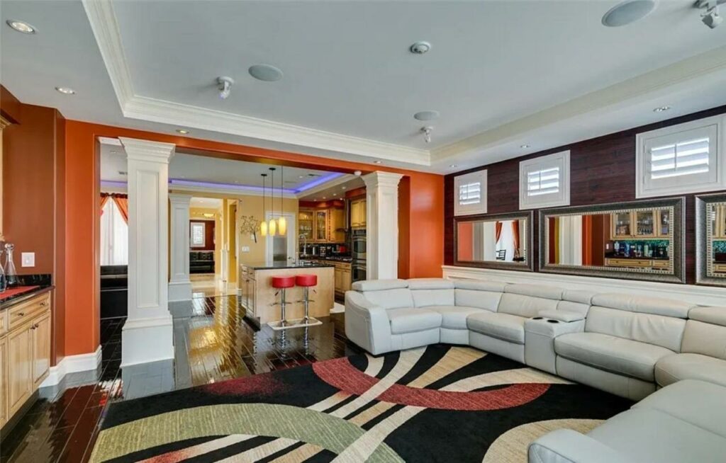 Sala de estar muito espaçosa na casa The Winner’s Retreat com dois sofaás brancos em L, um tapete preto com desenhos vermelhos, chão de madeira, indo adiante já é possível ver a cozinha
