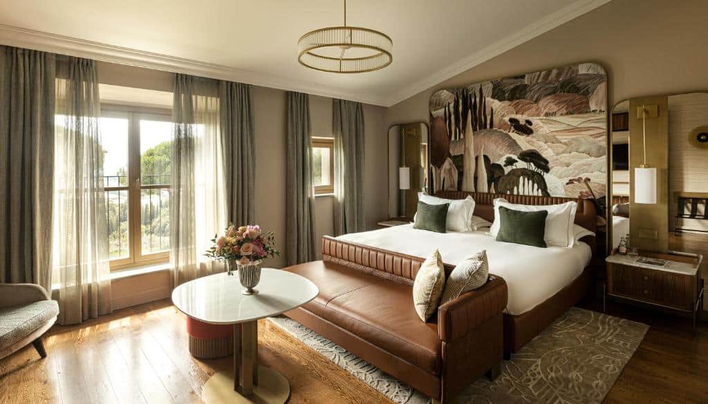 Quarto sofisticado do Toscana Resort Castelfalfi, usando cores no tom marrom e verde militar, com cama de casal, sofá de couro na frente, mesa com flor em cima e janelas grandes com vista da natureza