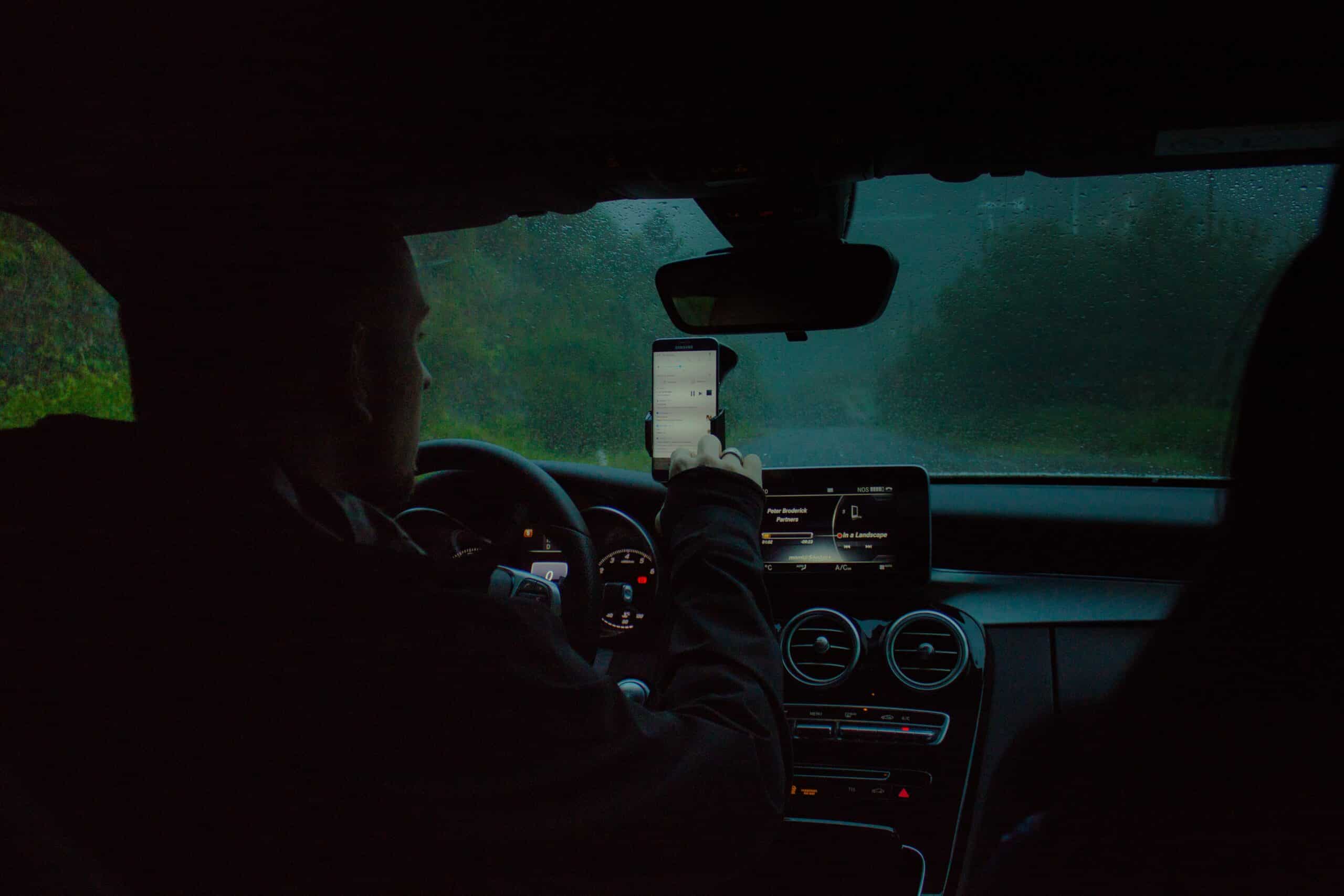 homem dirigindo um carro e mexendo no celular enquanto do lado de fora escurece e chove