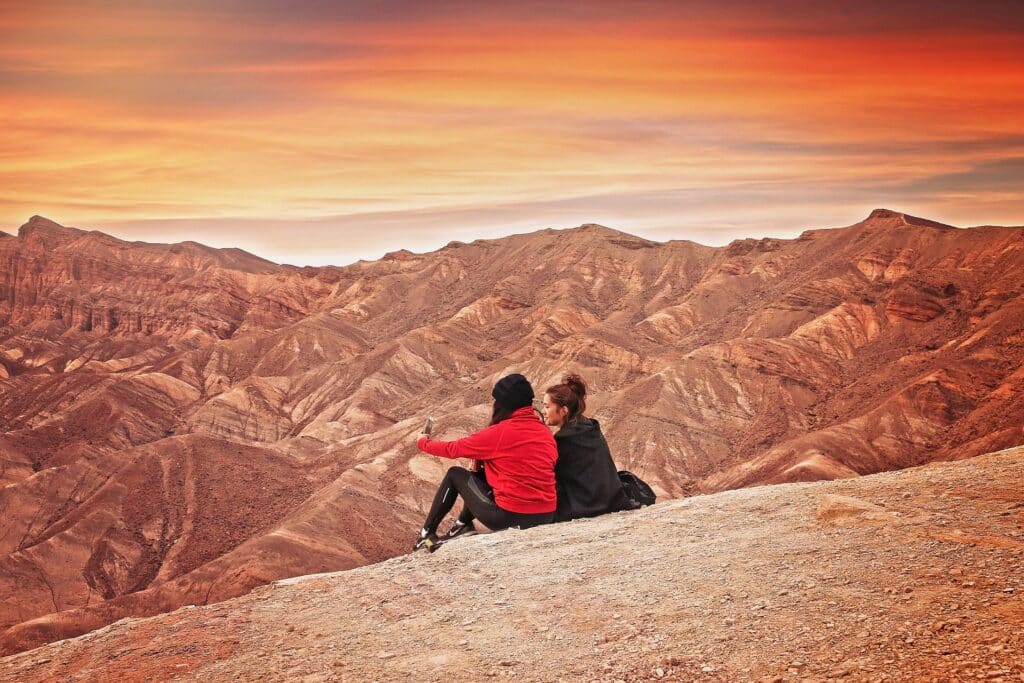 Duas mulheres tirando uma self no celular, sentadas no chão, no Vale da Morte, na Califórnia, com diversas montanhas alaranjadas em volta
