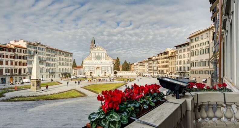 Varanda do Hotel Roma, com algumas flores vermelhas e vista da Praça Santa Maria Novella, com céu azul e várias nuvens brancas