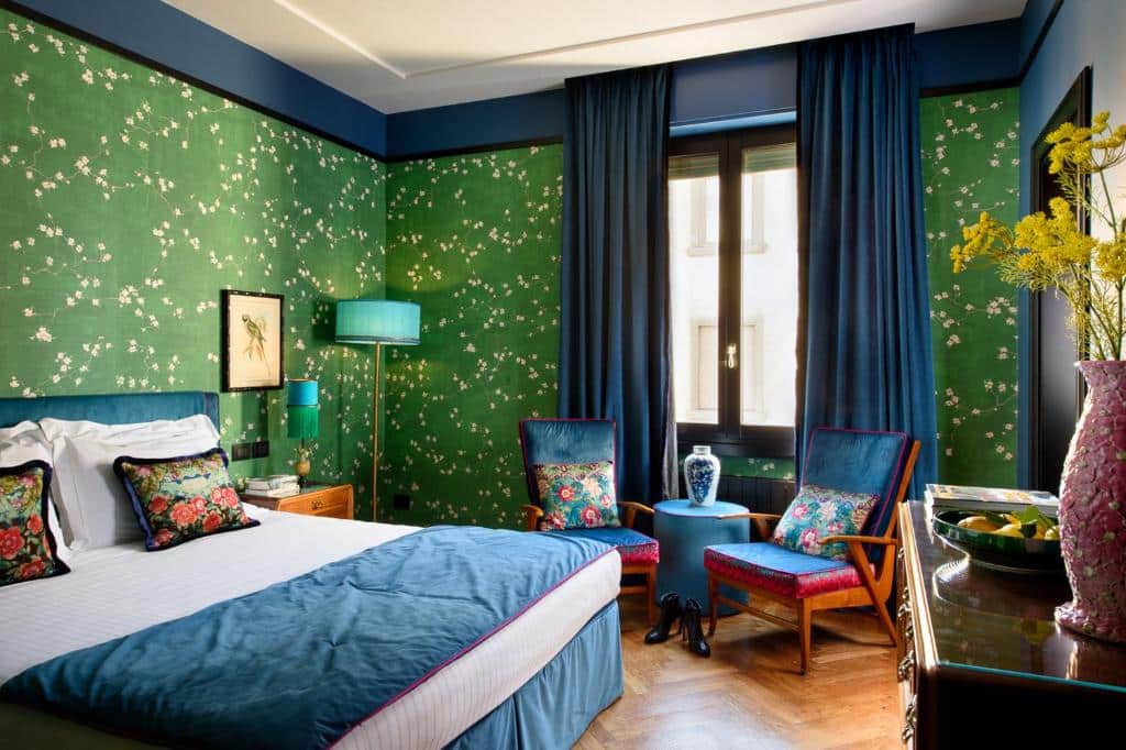 Quarto deluxe do Velona's Jungle Luxury Suites, com paredes verdes com desenhos de flores brancas, cortina azul turquesa, duas poltronas estofadas, cama de casal com almofadas e travesseiros em cima
