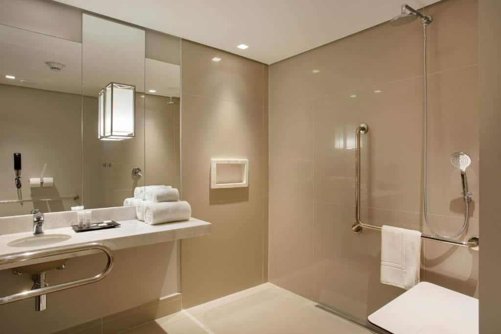 banheiro acessível do Venit Barra Hotel com espaço amplo, pia vazada com barras de apoio na bancada e dentro do boxe, onde há uma cadeira acessível e toalhas brancas disponíveis