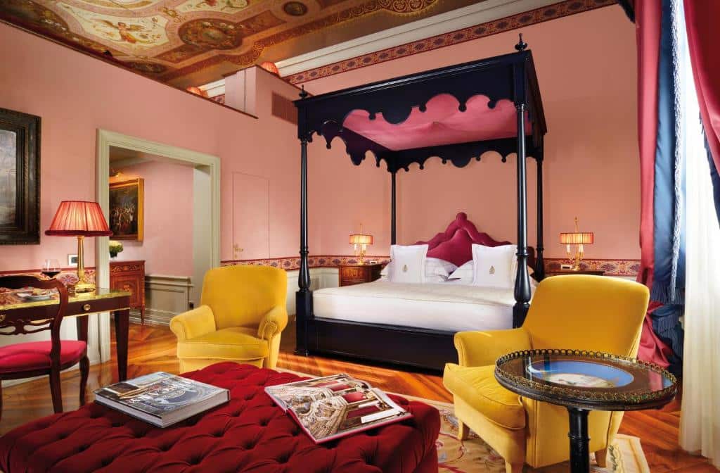 Quarto com decoração clássico do Villa Cora, com 80 m², destacando as cores vinho, rosa e amarelo, com duas poltronas  no tom mostarda, cama de casal grande, mesinhas de época com abajur aos lados e mesinha com uma cadeira estofada