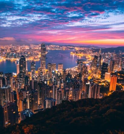 Vista aérea de prédios iluminados de Hong Kong vistos a noite, com céu alaranjado ao escurecer do dia.