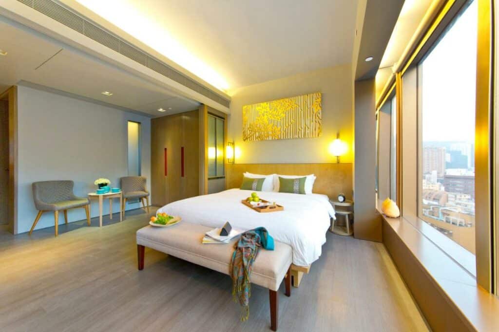 Quarta do hotel Wanchai 88 em Hong-Kong, possuindo uma cama ao centro com edredom branco e travesseiros, chão de assoalho, janela ao lado, duas poltronas e uma mesa perto da parede e abajures perto da cabeceira da cama.