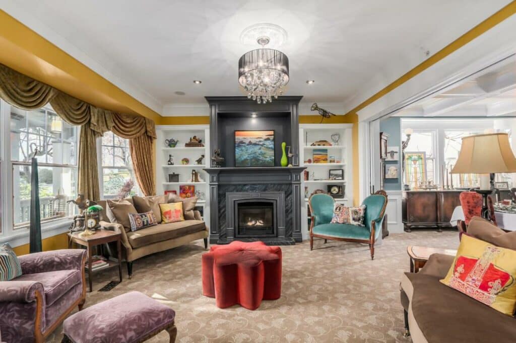Sala de estar ampla no West End Guest House com diversos sofás de cores diferentes como marrom e roxo, poltronas azuis e vermelhas, uma lareira preta com muitos enfeites nela, e do lado esquerdo, uma janela ampla com cortinas amarelo mostarda