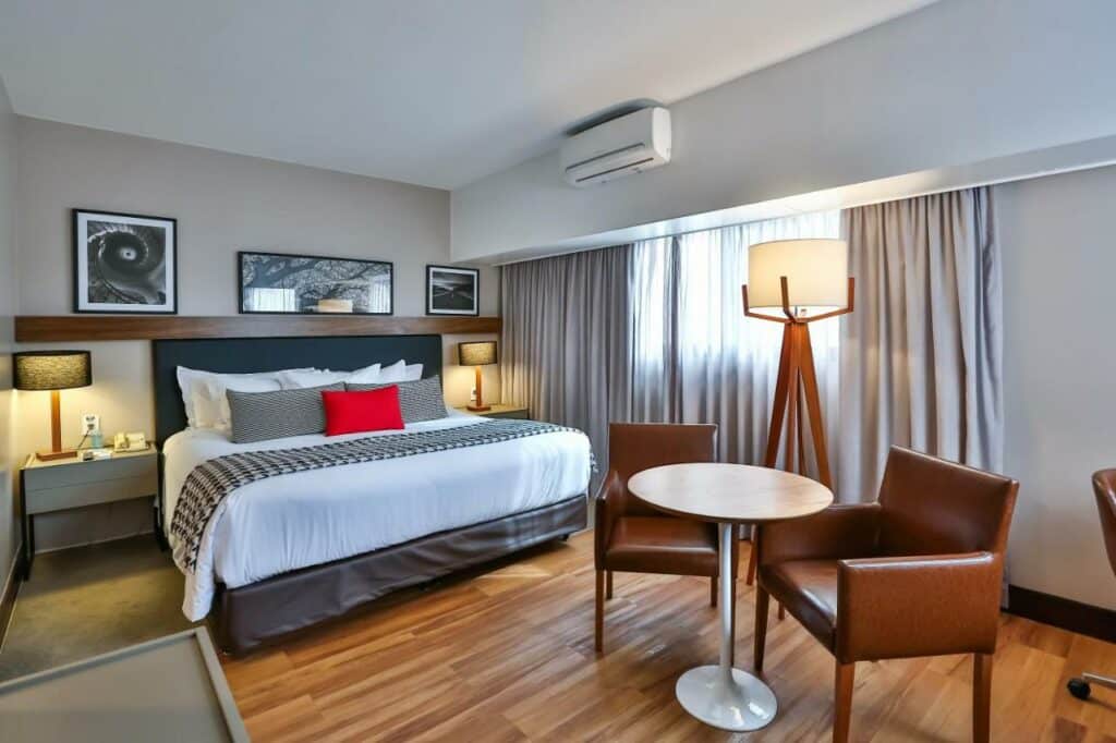 Quarto espaçoso do Wyndham São Paulo Berrini com uma cama de casal, uma janela com cortinas, um ar-condicionado, uma mesinha redonda pequena com duas poltronas de couro marrom, o chão é de madeira, para representar hotéis perto do Lollapalooza