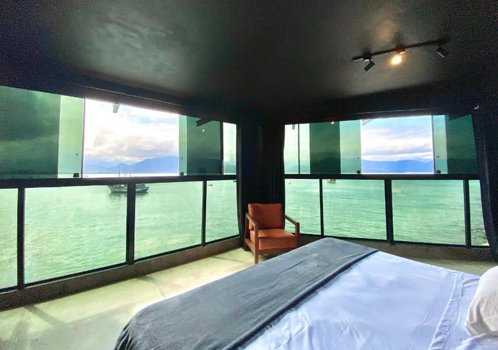 Quarto do 1819 Marine Hotel com uma cama de casal e um poltrona, o local é cercado por paredes e janelas de vidro com vista direta para o mar