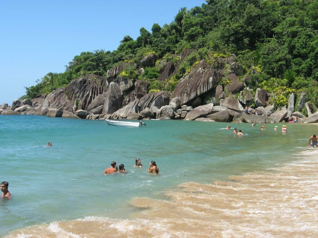 Uma praia em Ilhabela com rochas e muita mata perto do mar de águas claras com pessoas dentro