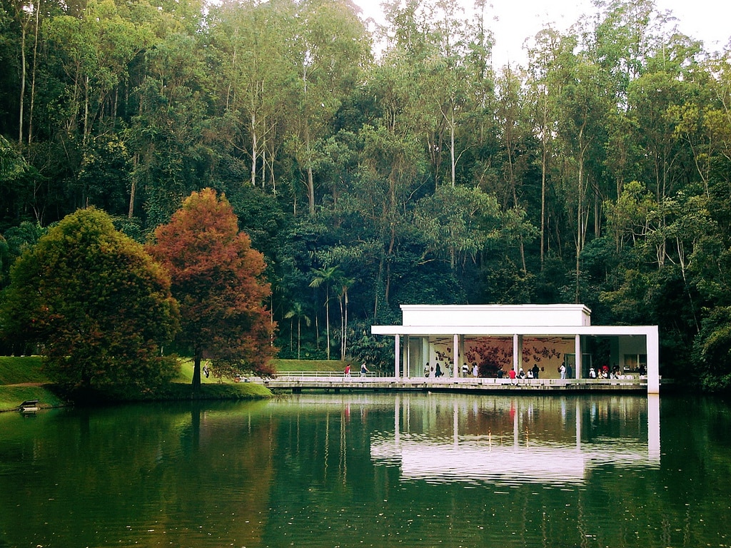 vista Instituto Inhotim, em Brumadinho, com um lago que está refletido árvores e a própria estrutura branca e aberta  e bem moderna que está além do lago