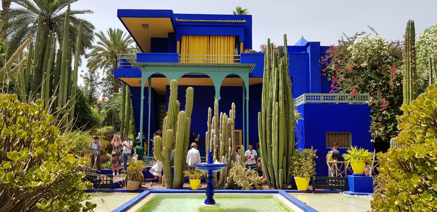 visitantes com celulares nas mães do Jardim Majorelle, com diversos cactos diferentes e altos próximo de uma fonte em uma piscina quadrada e um prédio atrás de um forte azul que contrasta com p verde dos cactos, que ilustra o post de chip celular Marrakesh