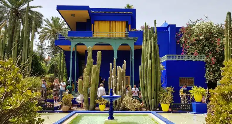 visitantes com celulares nas mães do Jardim Majorelle, com diversos cactos diferentes e altos próximo de uma fonte em uma piscina quadrada e um prédio atrás de um forte azul que contrasta com p verde dos cactos, que ilustra o post de chip celular Marrakesh