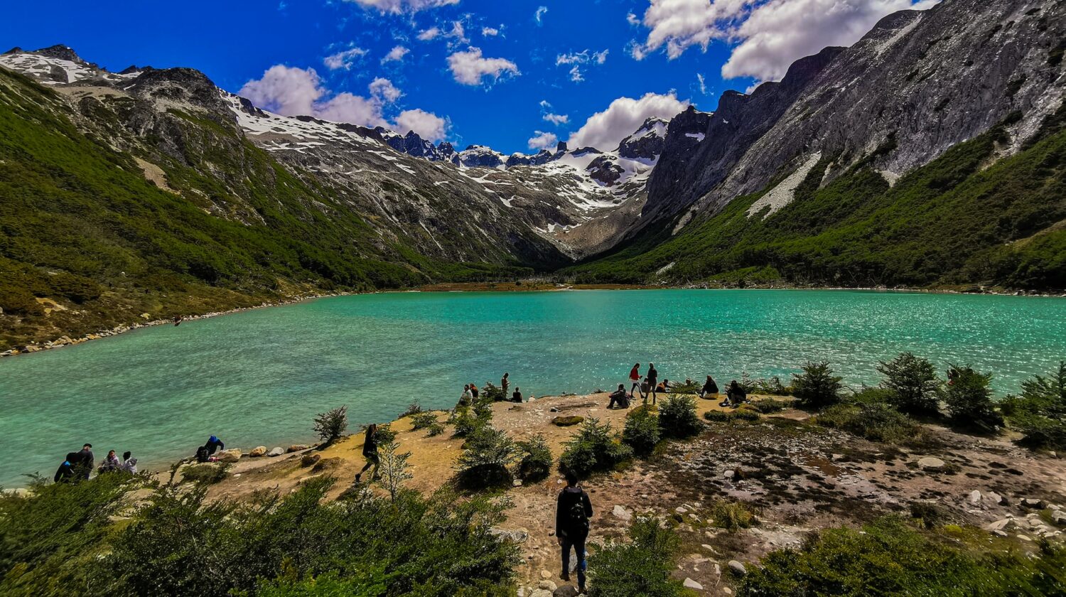 pessoas ao redor da Laguna Esmeralda, que ilustra o post de chip celular Ushuaia, estando em pé ou sentadas nas pedras baixas e vegetação escassa na beirada do lago esverdeado com montanhas ao redor e céu muito azul com poucas nuvens