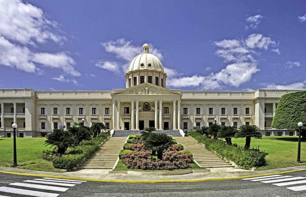 vista de frente do Palácio Nacional de Santo Domingo, na República Dominicana, com grama bem verde e flores, sendo uma estrutura clássica e imponente