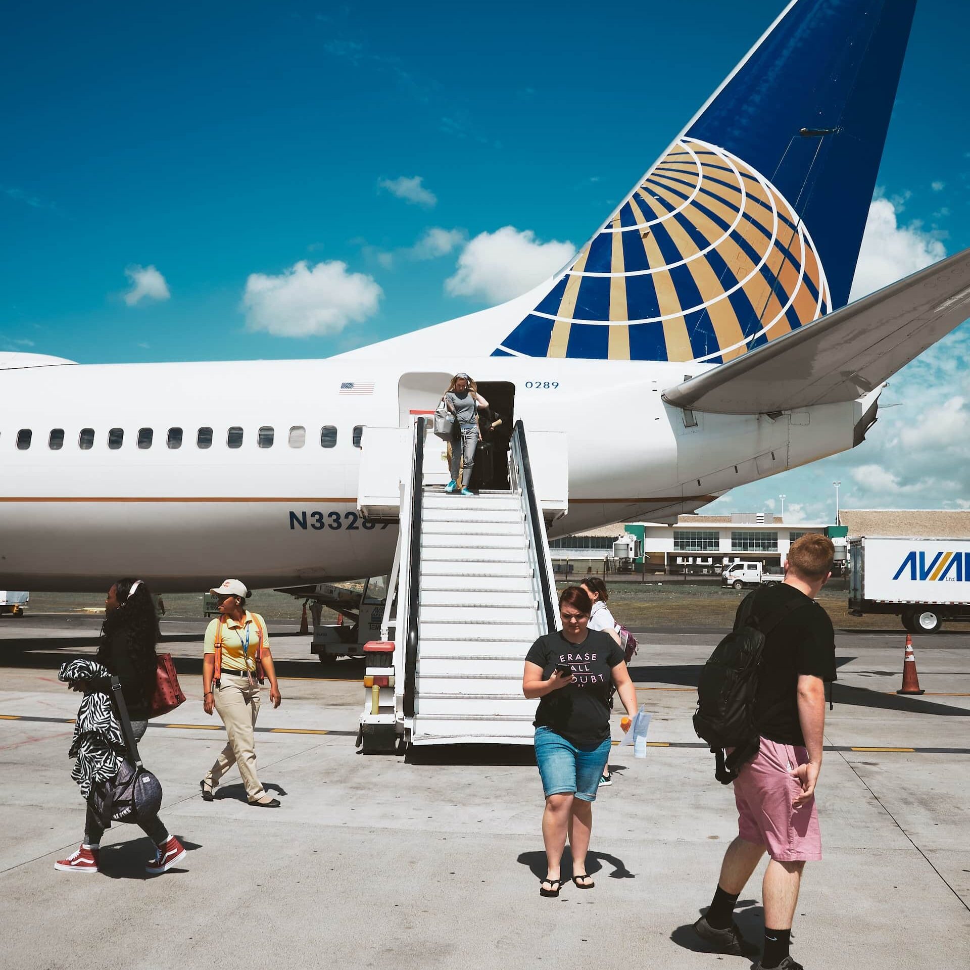 Pessoas andando na frente de um avião, que está com a escada dando acesso para os passageiros descerem do avião, sendo uma mulher com mala na mão descendo o degrau e outros andando em volta, e uma outra mulher de bermuda jeans e blusa preta mexendo no celular, ilustrando o chip de celular para Punta Cana