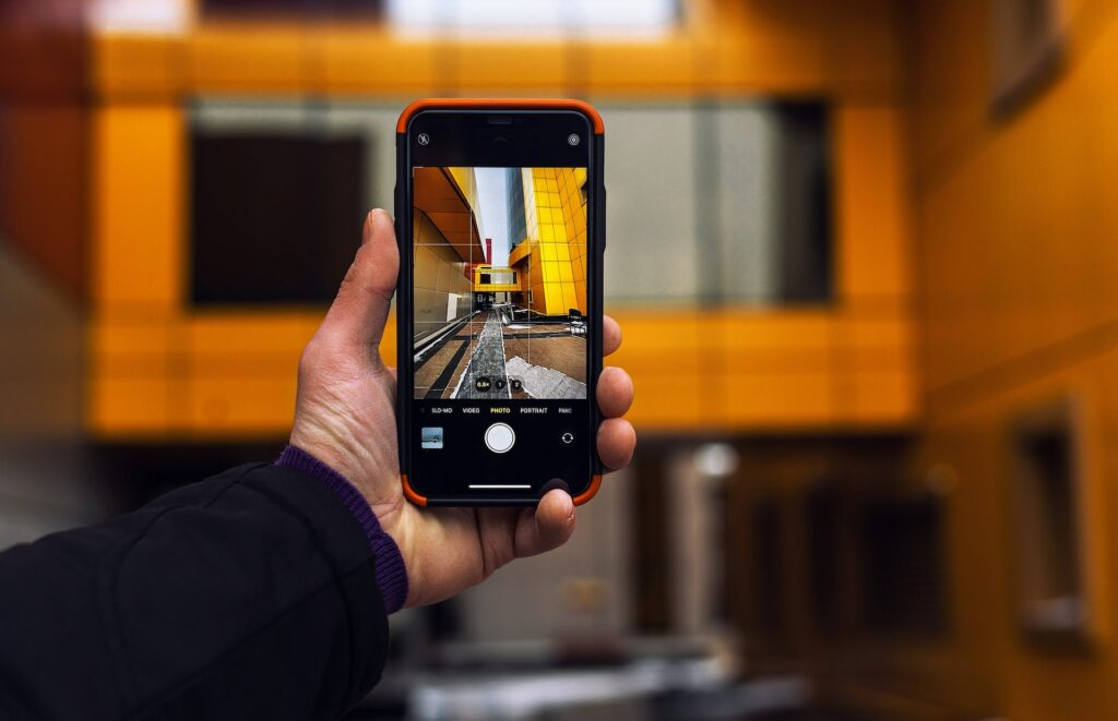 Uma mão branca masculina segurando um celular na vertical enquanto tira uma foto de um prédio amarelo com janelas grandes de vidro