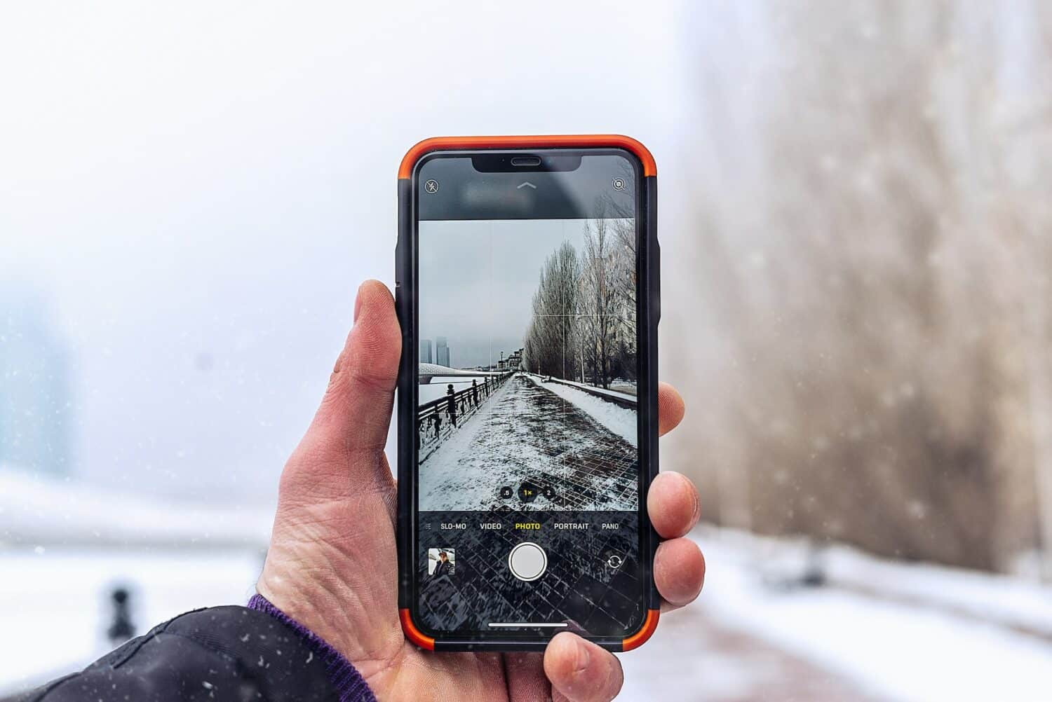 Uma mão branca masculina segurando um celular na vertical enquanto tira a foto de uma rua cheia de neve e com algumas árvores