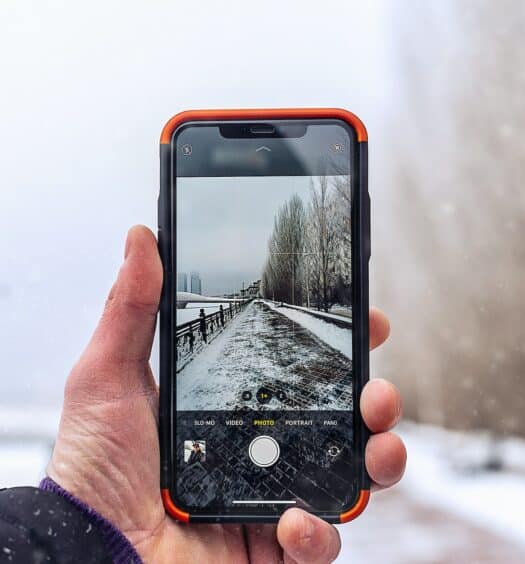Uma mão branca masculina segurando um celular na vertical enquanto tira a foto de uma rua cheia de neve e com algumas árvores