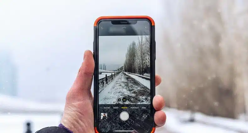 Uma mão branca masculina segurando um celular na vertical enquanto tira a foto de uma rua cheia de neve e com algumas árvores