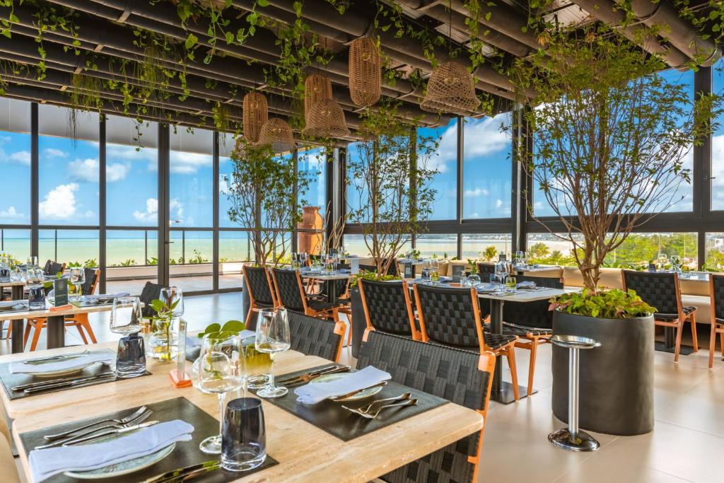 refeitório do BA'RA Hotel em João Pessoa com vista para a praia e o mar em um ambiente amplo, com bastante janelas e plantas. Há diversas mesas com quatro cadeiras, todas equipadas com louças elegantes