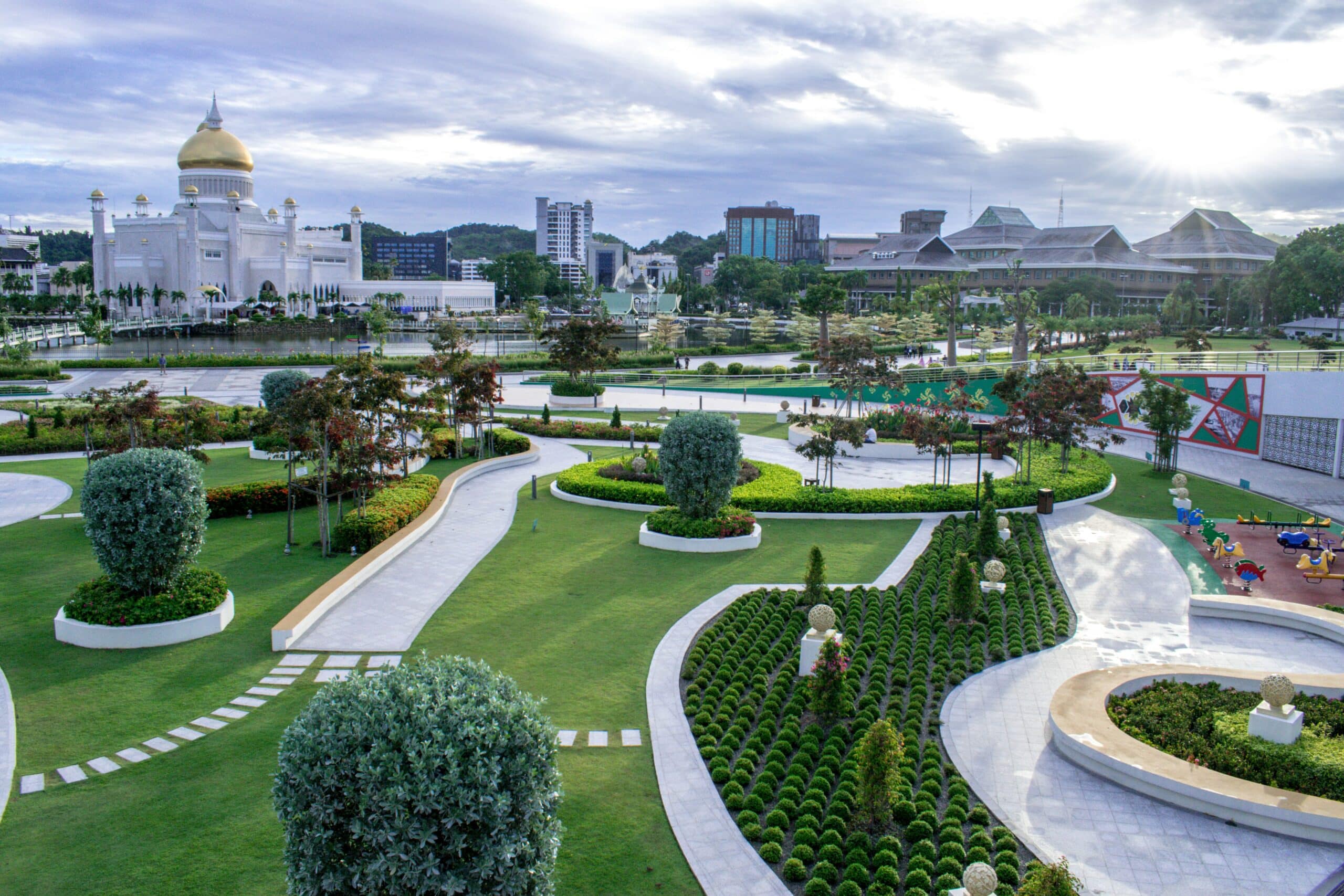 Vista da cidade de Bandar Seri Begawan, em Brunei, com um belo jardim a frente durante o dia e, ao fundo, tem um lago e em frente ao lago há uma enorme mesquita na cor branca, com teto dourado. Representa chip celular Brunei