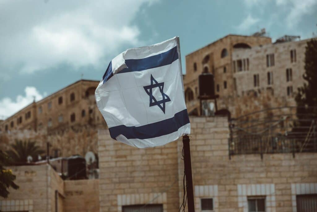 Bandeira de Israel tripudiando na região da cidade de Jerusalém, ao fundo algum edifício com paredes de tijolos cinzas para representar o seguro viagem Jerusalém.