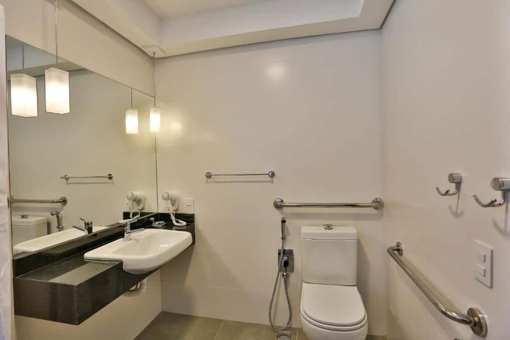 Banheiro do hotel Quality, em Blumenau, com barras de apoio em cima e no lado direito do vaso sanitário, e uma pia branca , embutida em um balcão de mármore preto, em uma altura mais baixa para hóspede com mobilidade reduzida