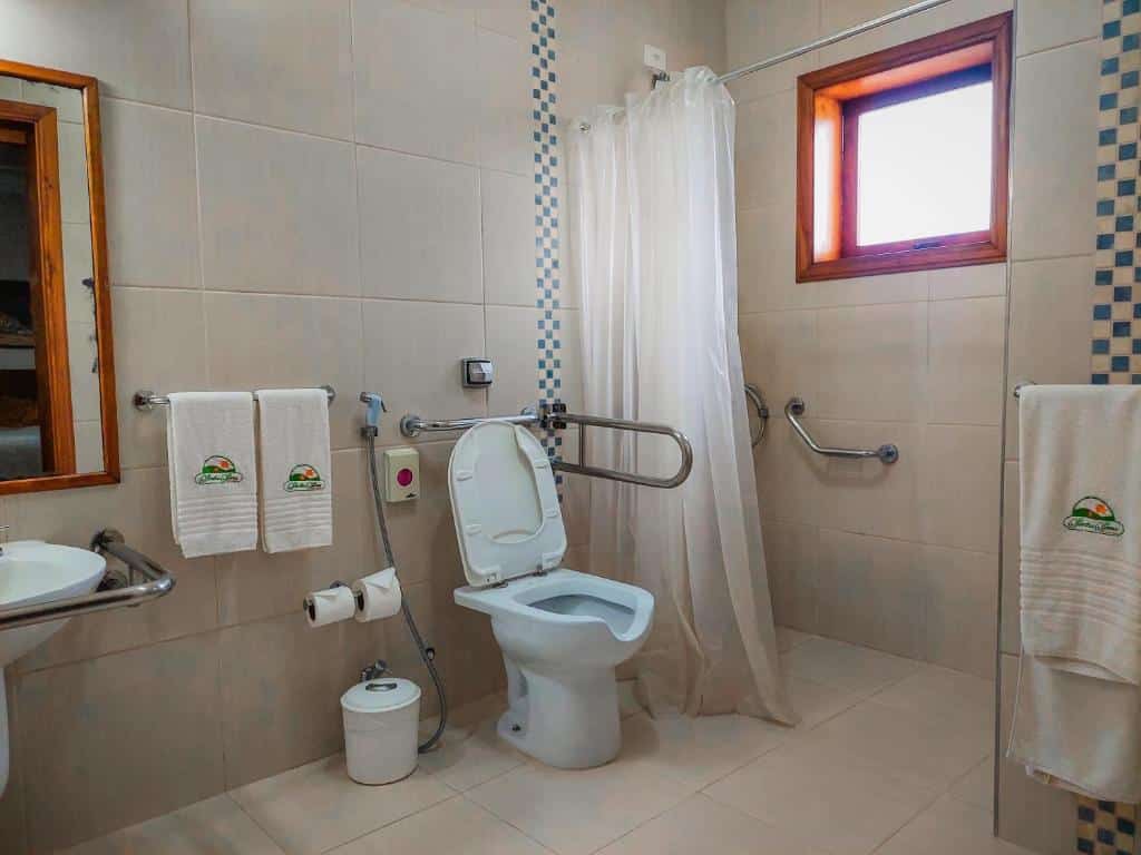 Banheiro para pessoas com mobilidade reduzida da Pousada Victoria Villa, na Serra da Mantiqueira, com um vaso (barras de apoio ao lado e em cima), uma pia (com barra de apoio na frente) e uma cortina de plástico na parte de banho com duas barras de apoio