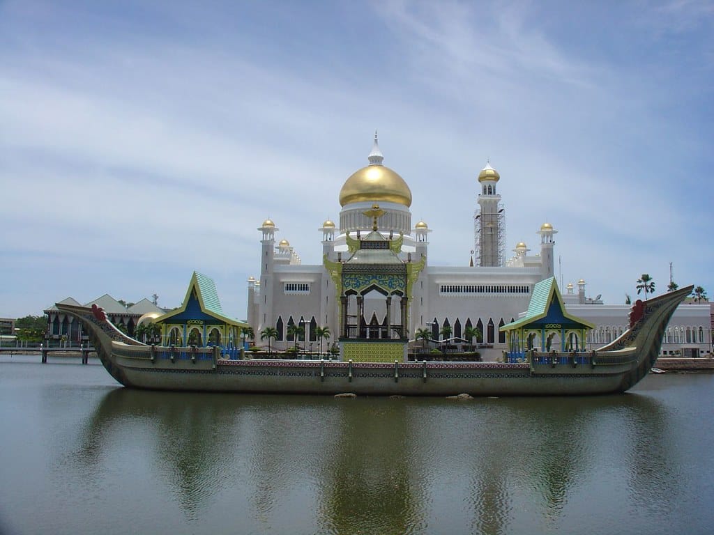 Barco no lago em frente da Grande Mesquita de Omar Ali Saifuddin, em Brunei, com a mesquita ao fundo de cor branca, teto dourado, durante o dia.