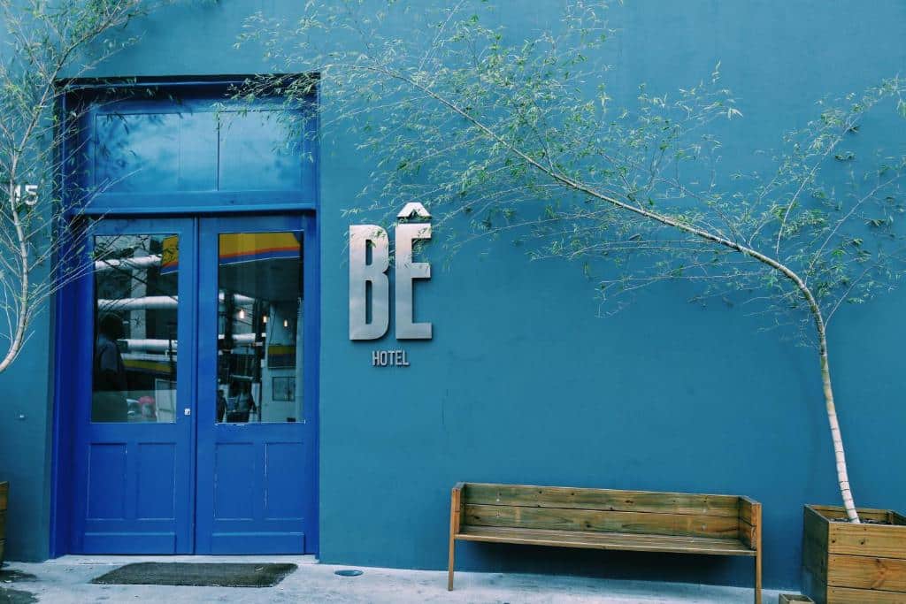 Fachada do Bê Hotel com uma parede azul, uma porta estilo antiga com vidro e o nome do hotel ao lado em prata, há também um banco de madeira e uma árvore do lado direito