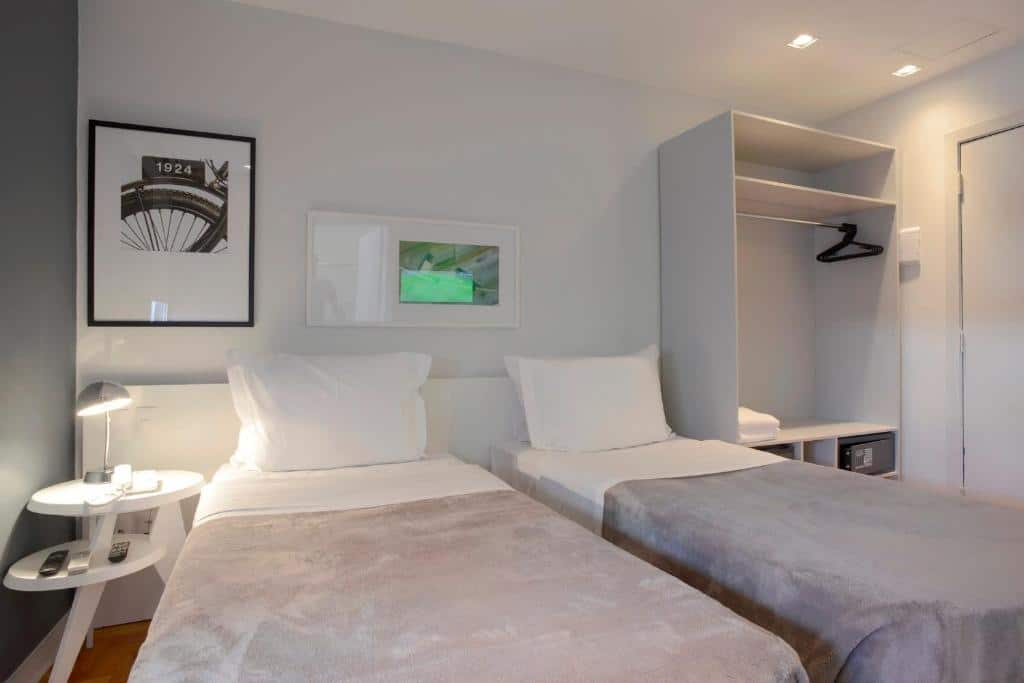Quarto do Bê Hotel com duas camas de solteiro, um armário de conceito aberto, e uma mesinha de cabeceira com uma luminária, para representar os hotéis perto do Allianz Parque