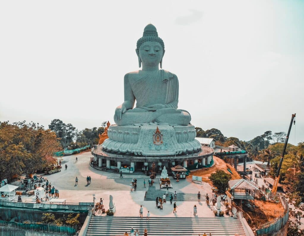 Estátua do Buda gigante no centro com uma escadaria logo a frente, algumas vegetações ao entorno e pessoas circulando em volta do local para representar o seguro viagem Phuket.