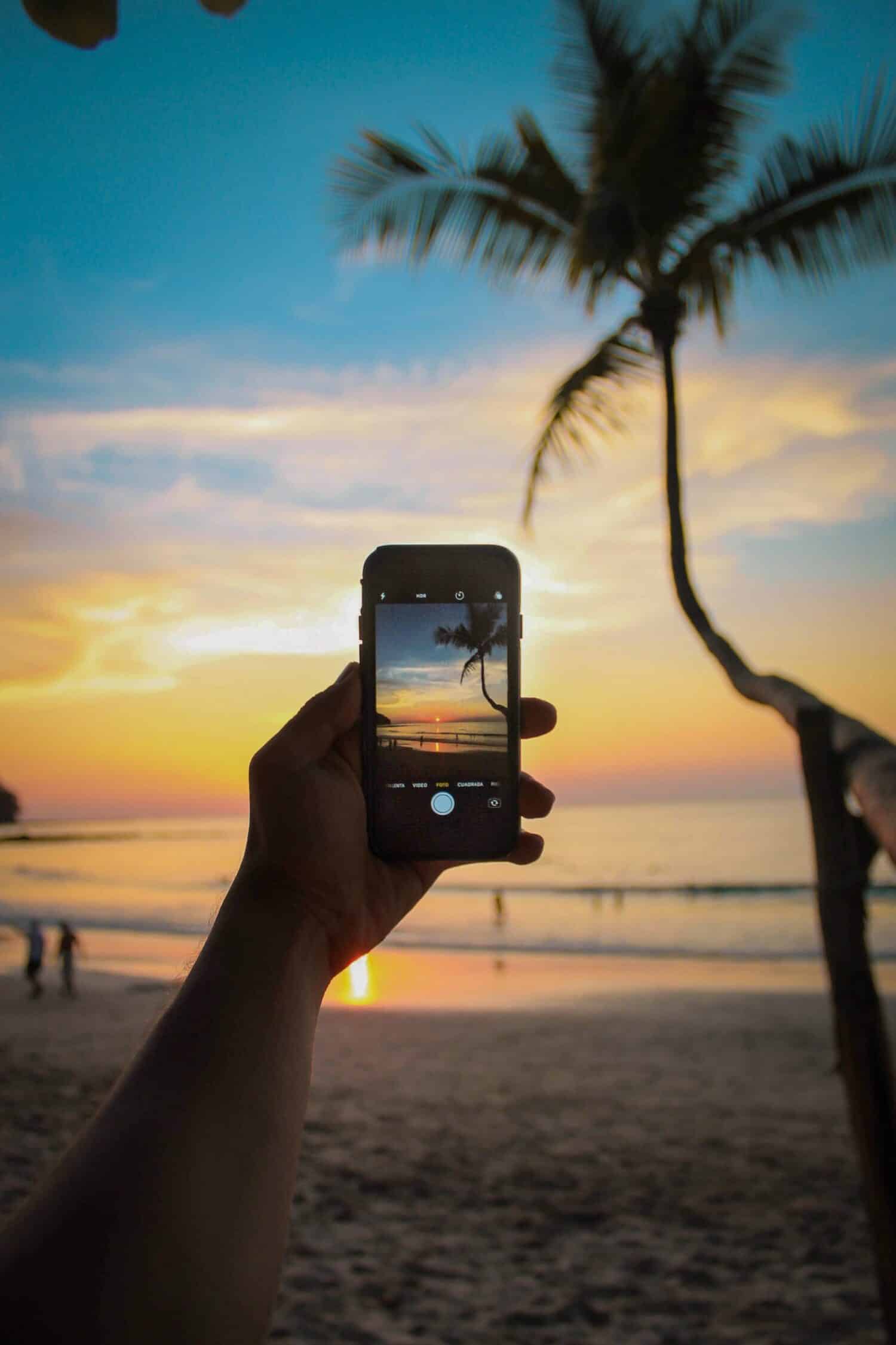 Uma mão masculina segurando um celular na vertical enquanto tira uma foto de uma praia no entardecer