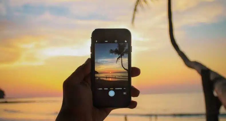 Uma mão masculina segurando um celular na vertical enquanto tira uma foto de uma praia no entardecer