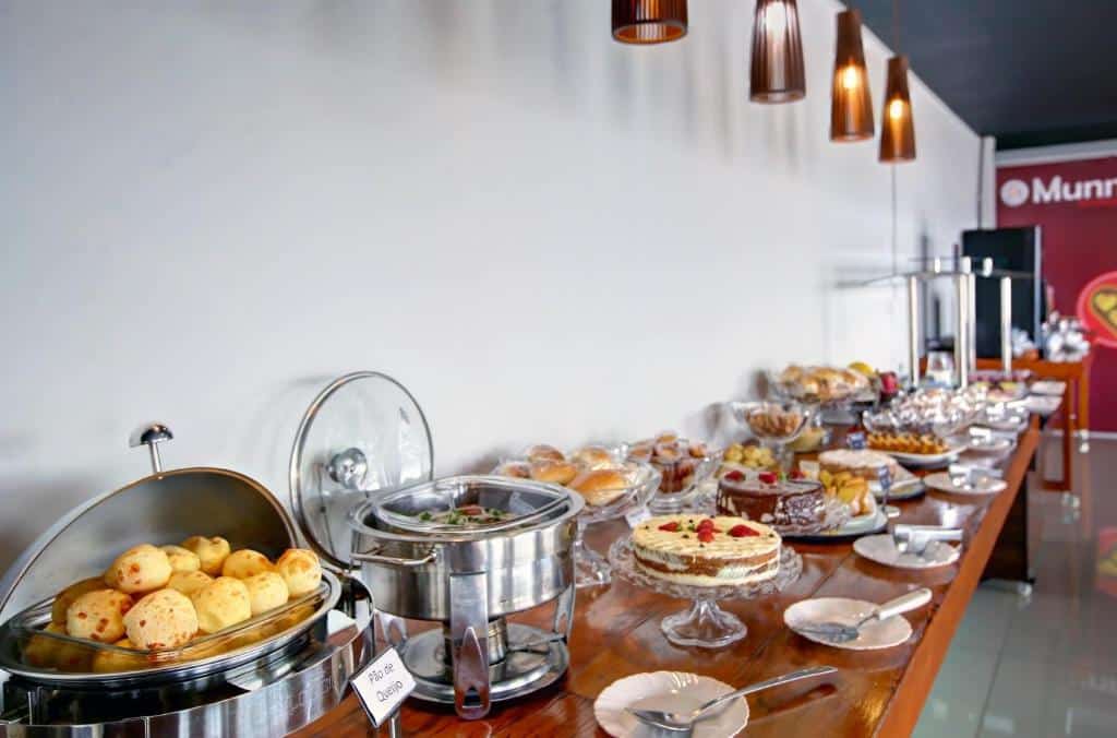 buffet de café da manhã no Go Inn Betim - MG, uma das pousadas em Betim, com diversas opções incluindo pães de queijo, tortas, bolos, salgadinhos e muito mais, com luminárias acima e máquina de café mais ao fundo