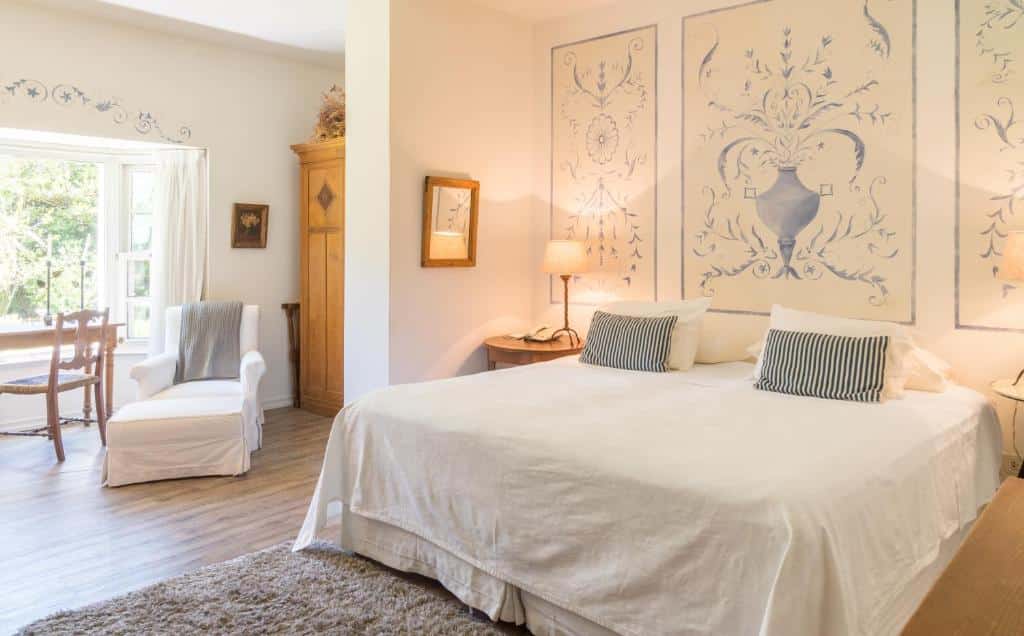Quarto do Hotel L'Auberge com cama de casal, cômoda ao lado da cama com luminária e poltrona branca do lado esquerdo.