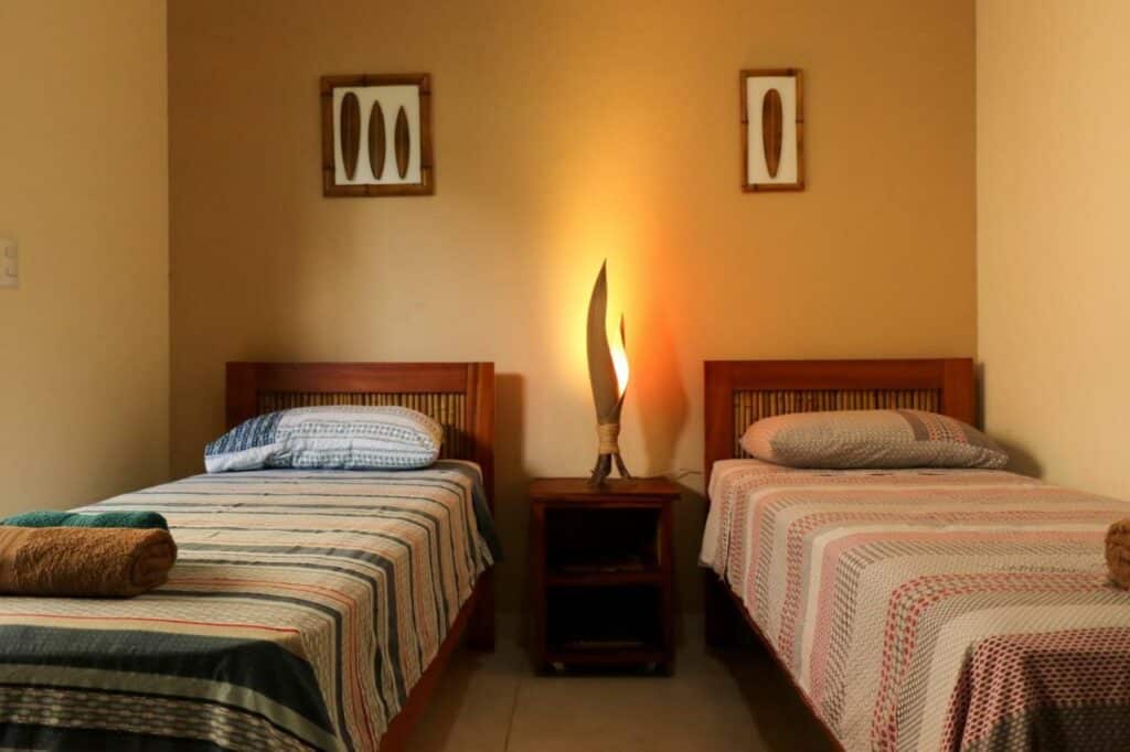 Quarto no Casa Viva Itamambuca com duas camas de solteiro, com travesseiros e toalhas sob as camas, e entre as duas há uma pequena mesinha de cabeceira com um abajur
