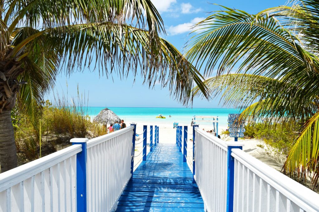 Deck de maneira azul com as laterais brancas, dando acesso a uma praia de Cuba, com água azul claro, areia branca e algumas pessoas aproveitando o sol num dia ensolarado