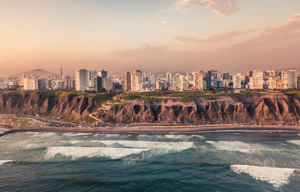 vista panorâmica da costa de Miraflores com ondas batendo na praia e uma costa alta em que se vê a cidade com vários prédios ao pôr do sol, para ilustrar o post de chip celular Lima