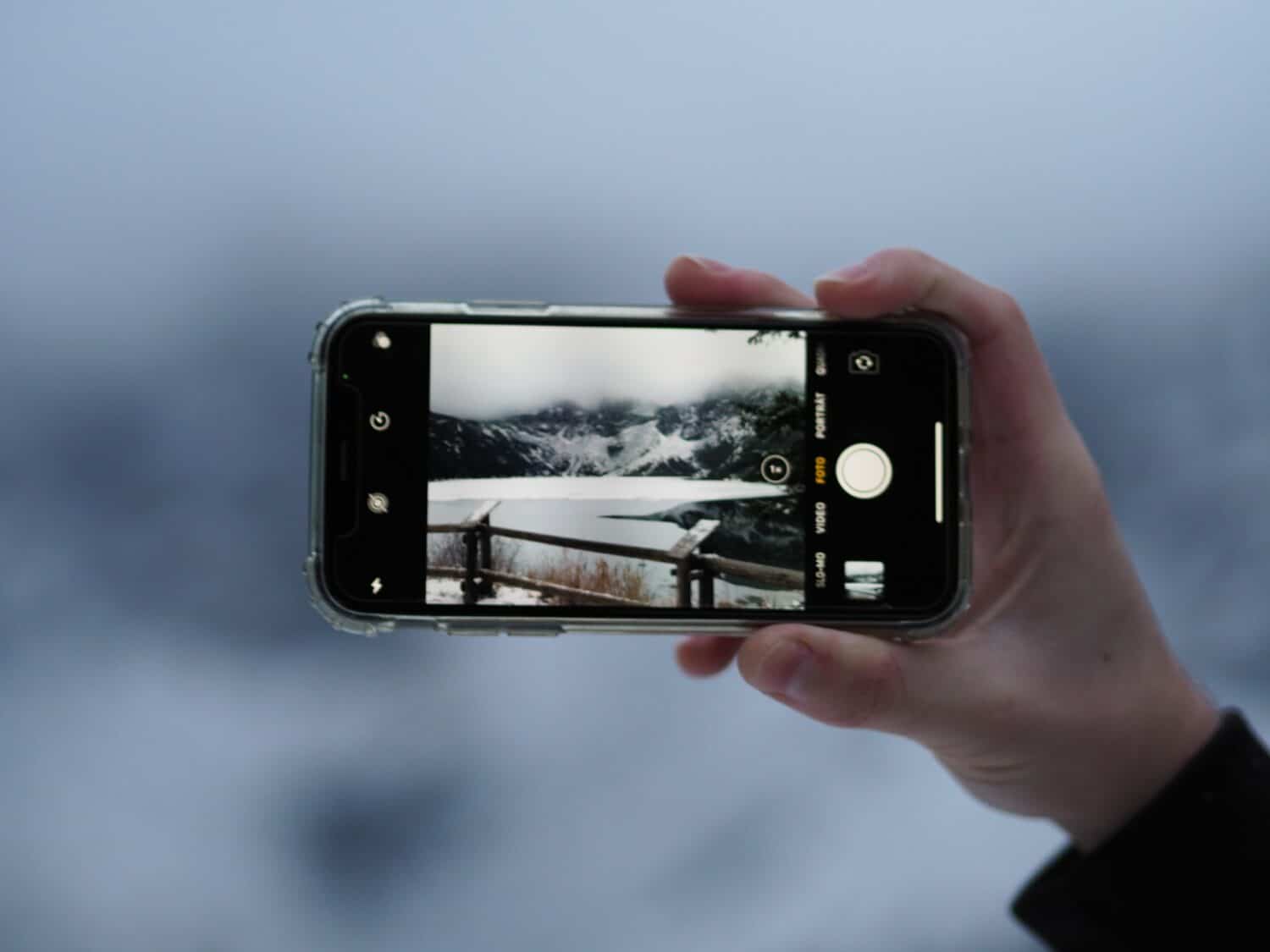 Mão branca segurando o celular e fotogrando uma região da Cracócia, composta por neve, montanha e uma pequena ponte de madeira